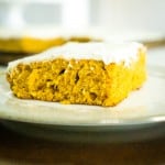slice of pumpkin cake with corner facing out. thin layer of cream cheese frosting on top, on slilver and white dessert plate