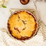 plum cobbler on marble counter with blue and white tea towel, fresh plum, and white flower nearby
