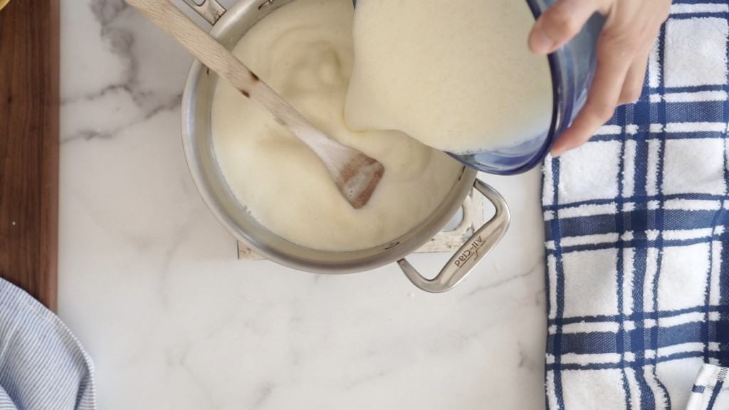 blue glass bowl with egg, milk, and cream being poured into saucepan with milk and cream