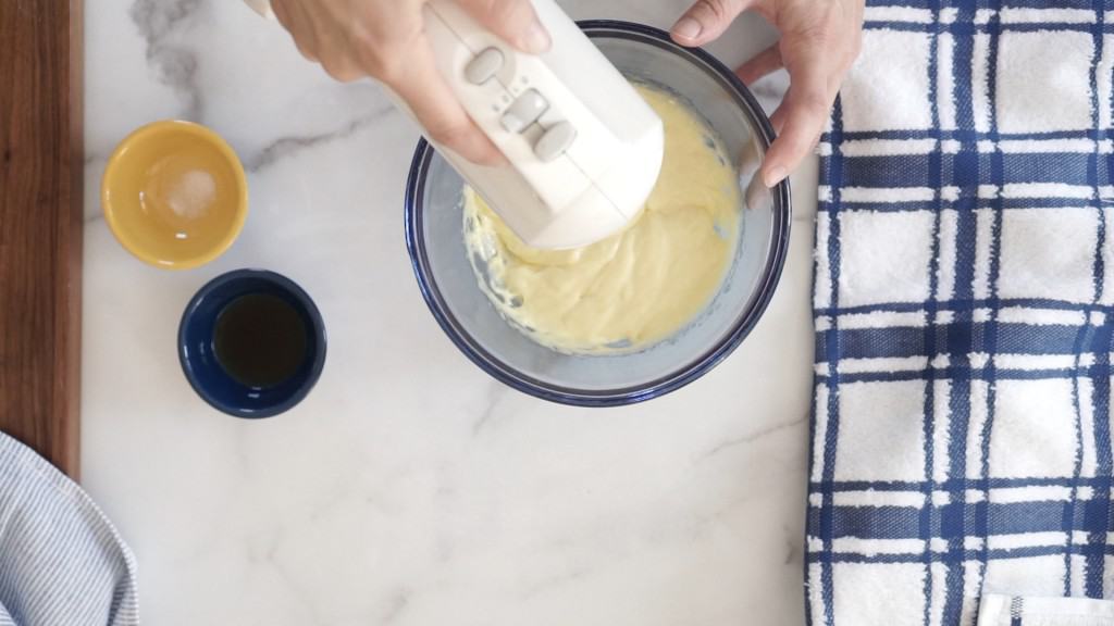 hand mixer beating egg yolk and sugar in blur glass bowl.