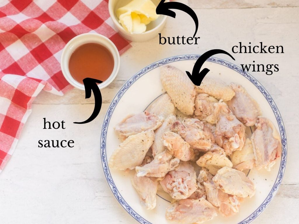 raw chicken wings on plate next to small container of hot sauce and butter