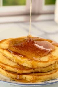 A stack of fluffy sourdough pancakes on a plate, topped with a drizzle of maple syrup, with syrup cascading down the sides of the pancakes.