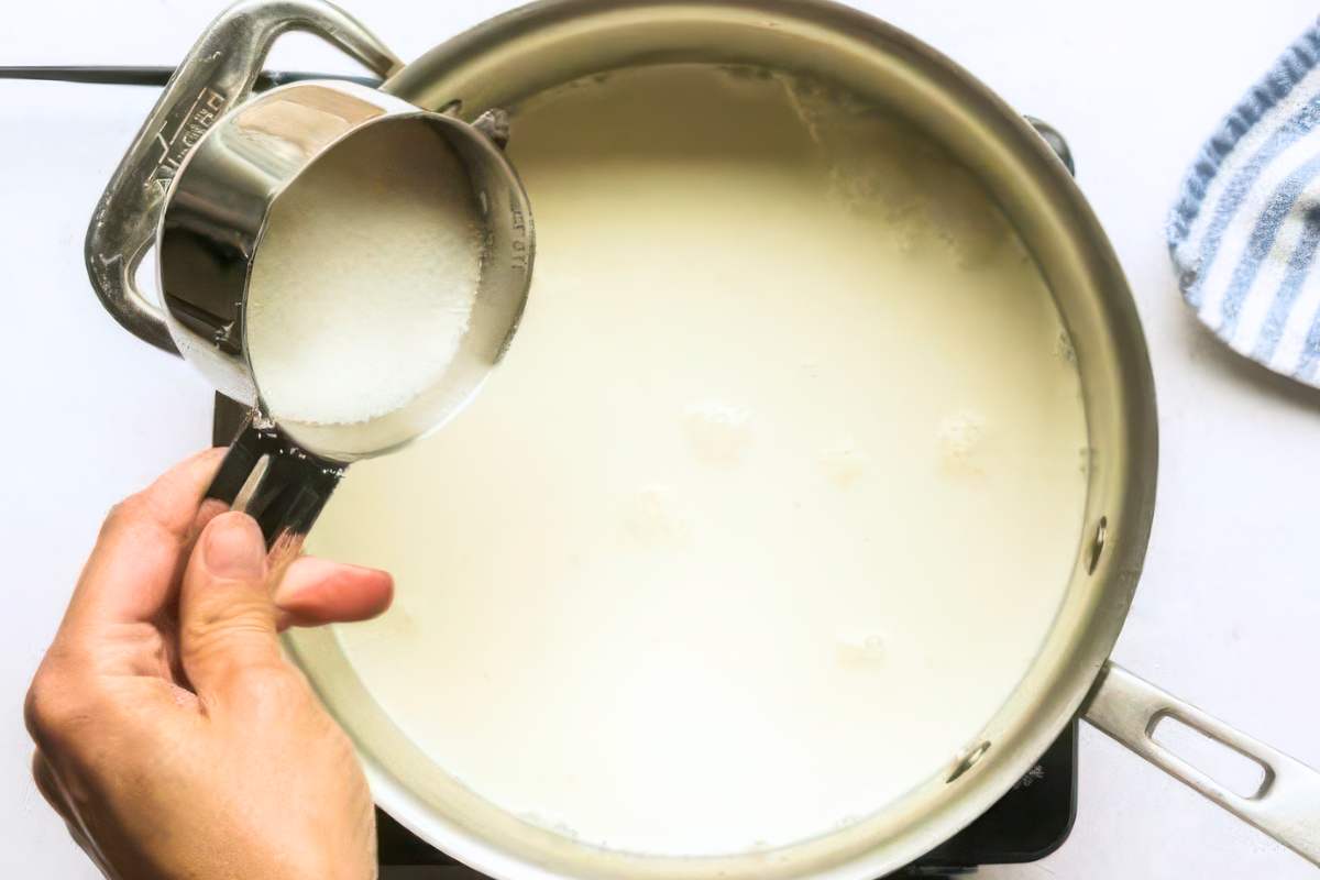 Adding sugar to milk and cream mixture in a saucepan on stovetop