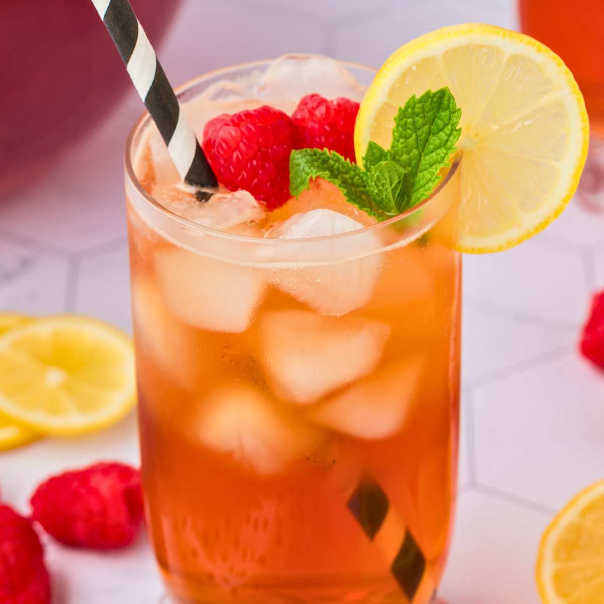 glass of raspberry peach tea with ice cubes and straw, garnished with raspberries, lemon, and mint.