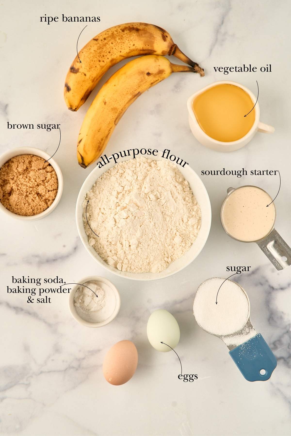 Overhead view of sourdough banana cake ingredients labeled and arranged on a marble surface.