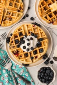A top-down view of a stack of buttermilk blueberry waffles topped with whipped cream and fresh blueberries, surrounded by extra blueberries and forks.