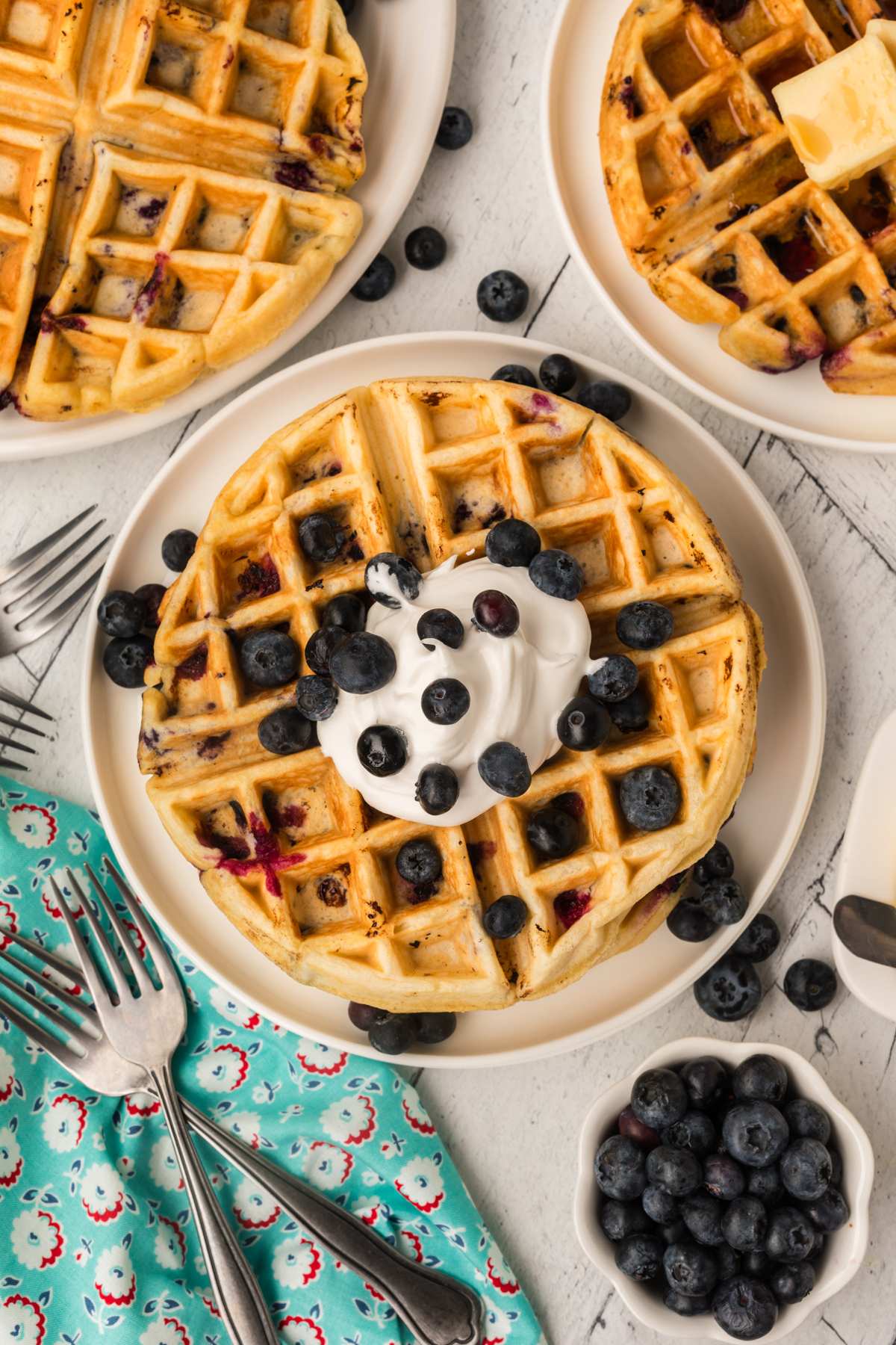 A top-down view of a stack of buttermilk blueberry waffles topped with whipped cream and fresh blueberries, surrounded by extra blueberries and forks.