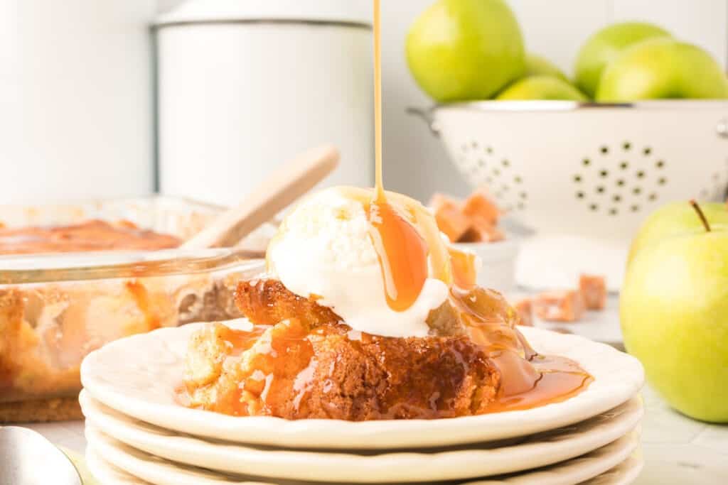 A slice of apple cobbler topped with a scoop of vanilla ice cream and drizzled with caramel sauce on a plate. In the background, a baking dish of cobbler, a white colander with green apples, and some caramel cubes are visible.