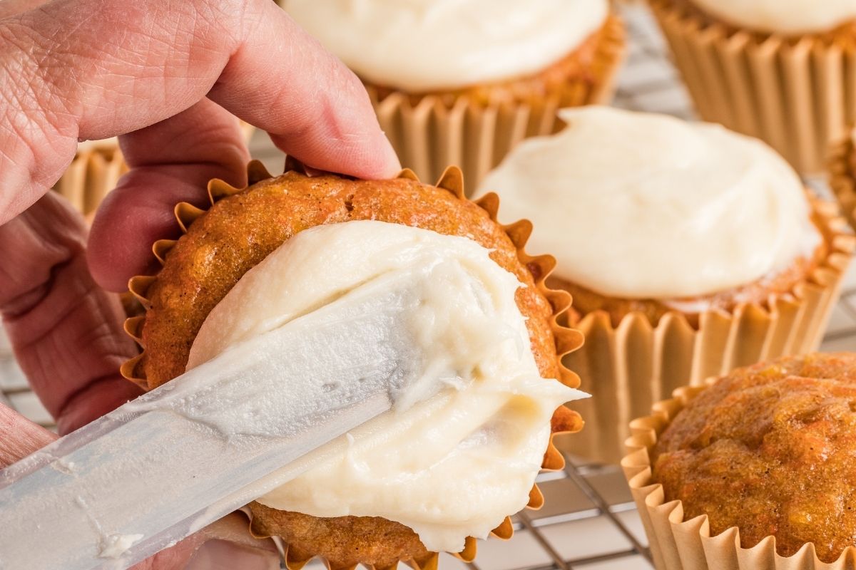 Hand spreading cream cheese frosting on a carrot cupcake with a knife.