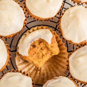 Frosted carrot cupcakes on cooling rack with one bitten to show inside.