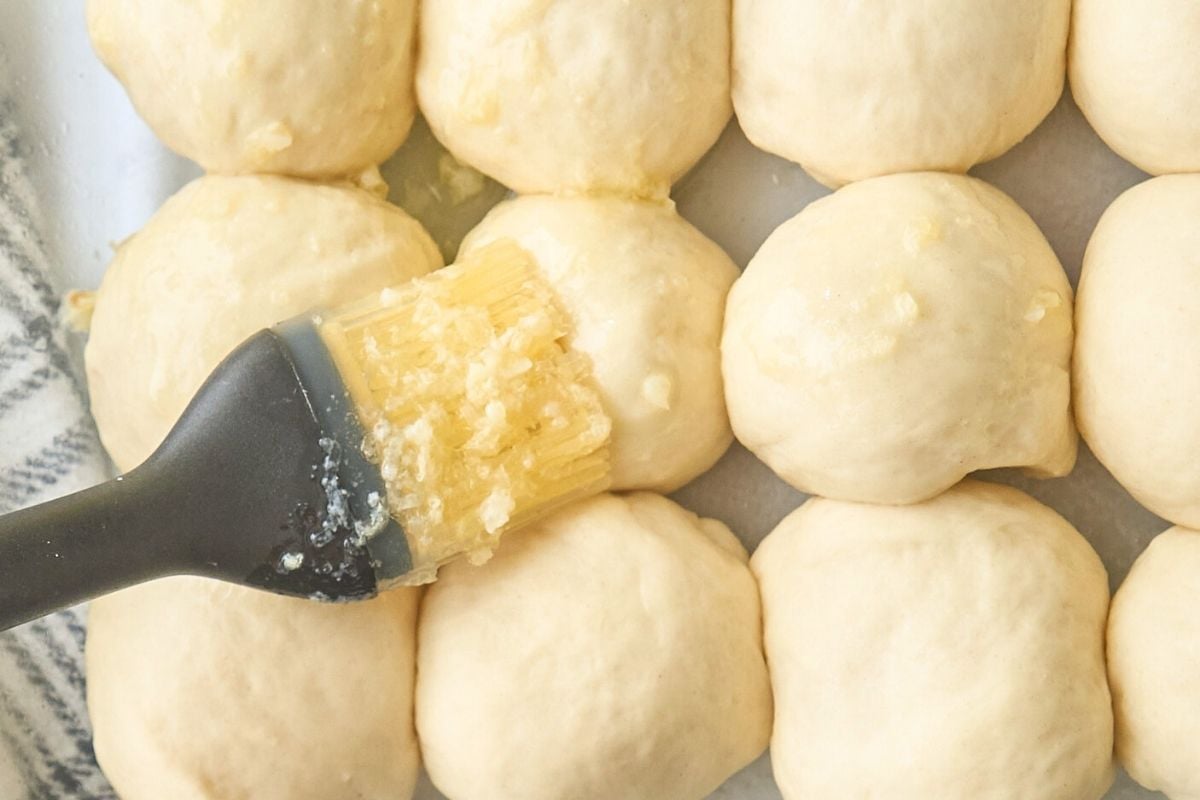 Pastry brush applying garlic butter mixture to risen dinner rolls in baking pan.