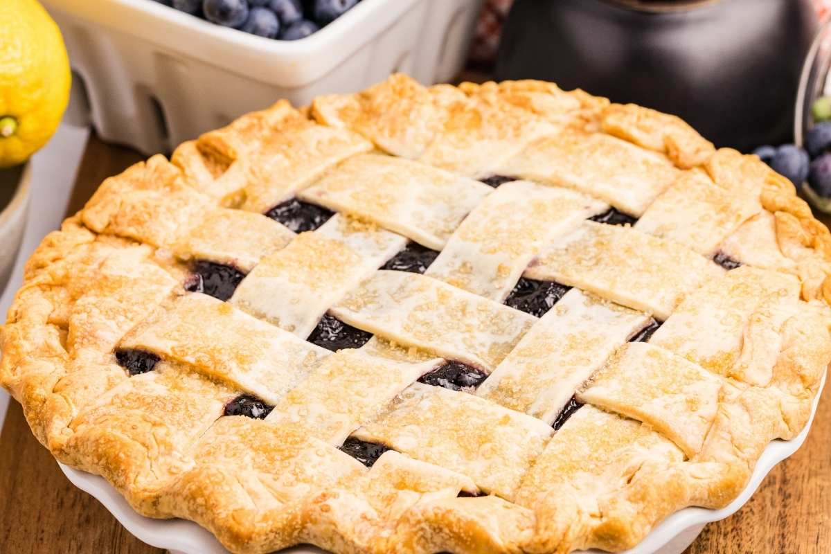 Golden lattice-topped blueberry pie with a lemon and berries nearby.
