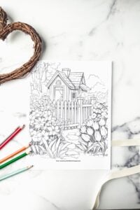 cottage coloring page on white marble with colored pencils and twine wreath nearby.