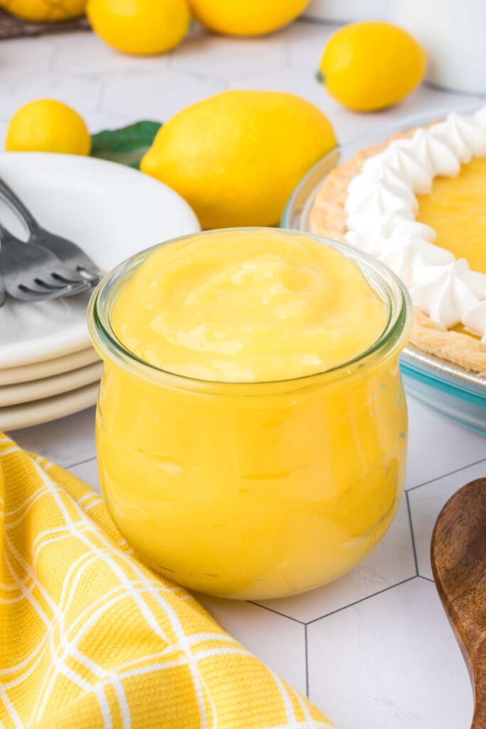 A clear glass bowl of creamy lemon pie filling on a kitchen counter, with a pie topped with whipped cream in the background, alongside a yellow and white checkered cloth and a wooden heart-shaped spoon.