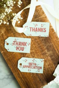 A selection of appreciation tags laid out on a wooden surface, with designs saying 'THANKS', 'THANK YOU', and 'WITH APPRECIATION' in red, accented with small floral details.
