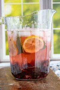 A side view of a full pitcher of blackberry lemon infused water, with layers of lemon slices, mint leaves, and blackberries at the bottom, placed on a wooden table with a green outdoor background visible through the window.