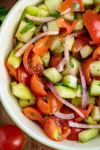 Close-up of cucumber tomato salad in a bowl, showing the mix of diced cucumbers, quartered tomatoes, and slices of red onion, seasoned with herbs.