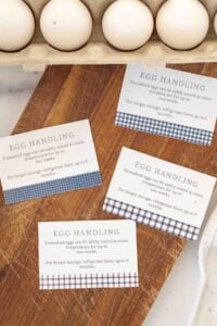A rustic wooden kitchen countertop with a carton of fresh eggs and printable egg handling instruction cards scattered around, featuring a blue gingham and stripe pattern."