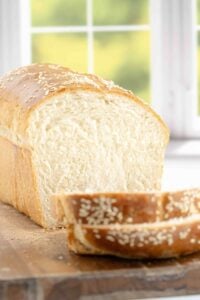 A loaf of Italian sandwich bread on a cutting board, with a few slices cut, showcasing the soft, airy texture inside and sesame seed crust.