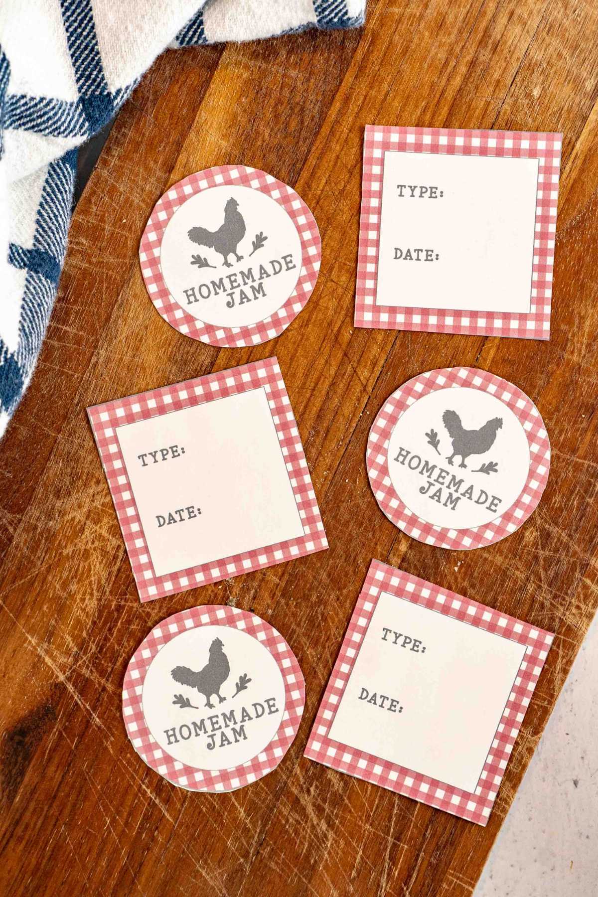 A rustic wooden tabletop displaying an assortment of printable jam jar labels, both round and rectangular, featuring a red and white gingham pattern and a chicken silhouette for homemade jam