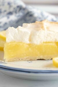 Side view of a slice of lemon meringue pie on a plate, showing the defined layers and texture, with lemon slices and a blue-patterned cloth in the background.