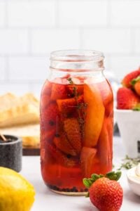 The jar of pickled strawberries takes center stage on a kitchen table complete with a striped cloth, surrounded by ingredients and tools used in the recipe, suggesting a warm, homemade culinary experience.