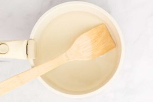 A wooden spoon stirs the pickling liquid in a cream-colored saucepan, emphasizing the hands-on aspect of preparing the recipe.
