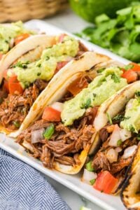 Three soft tacos filled with shredded beef, topped with guacamole, diced tomatoes, and onions on a white plate with a lime wedge on the side.
