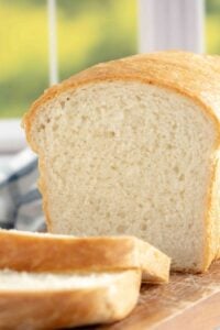 A close-up of a single loaf of bread, cut open with several slices laid out in front. The bread has a golden crust and a soft, airy interior, showcased in a bright, homey kitchen setting.
