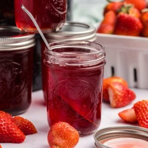 a jar of homemade strawberry syrup with a metal spoon inside with strawberries around.