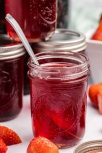 A jar of homemade strawberry syrup with a metal spoon inside, fresh strawberries and sealed jars in the background on a white surface.