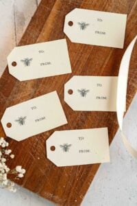 "A set of four printable 'to and from' gift tags laid out on a wooden surface, each featuring a detailed bee illustration above the words 'to' and 'from' in a classic serif font."