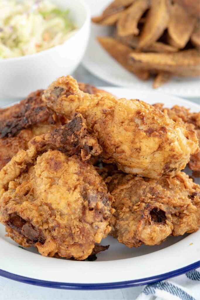 Golden brown fried chicken piled on a white plate with blue trim, served alongside a side dish of coleslaw and potato fries in the background.