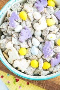 A colorful bowl of Easter Muddy Buddies, featuring Rice Chex cereal pieces coated in powdered sugar, with pastel-colored frosted animal crackers, yellow and white candy eggs, and a vibrant mix of Easter-themed candies, displayed in a blue bowl with a patterned rim, set against a yellow patterned background with small red flowers.
