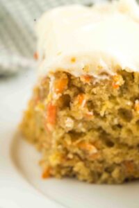Close-up of a slice of sourdough carrot cake on a plate, showing a moist crumb with visible flecks of grated carrot and raisins, topped with a smooth layer of cream cheese frosting.