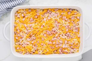Same casserole dish showing a layer of diced ham added on top of the cheese and hash browns.