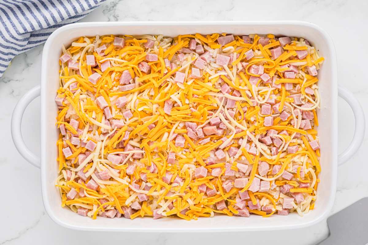 Same casserole dish showing a layer of diced ham added on top of the cheese and hash browns.
