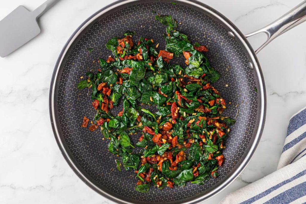 Sautéed spinach and sun-dried tomatoes in a skillet, prepared for adding to a Tuscan chicken dish.