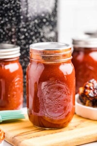 A sealed pint-sized mason jar filled with homemade smoky barbecue sauce, set on a wooden surface with blurred jars and barbecue in the background.