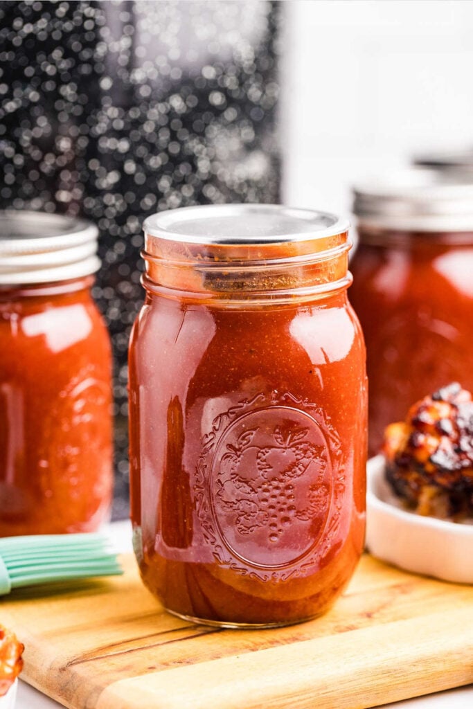 A sealed pint-sized mason jar filled with homemade smoky barbecue sauce, set on a wooden surface with blurred jars and barbecue in the background.