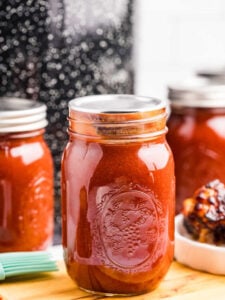 A sealed pint-sized mason jar filled with homemade smoky barbecue sauce, set on a wooden surface with blurred jars and barbecue in the background.