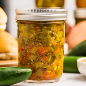 A clear glass jar filled with colorful jalapeño relish in sharp focus, with whole jalapeños, buns, and cornmeal in the blurred background