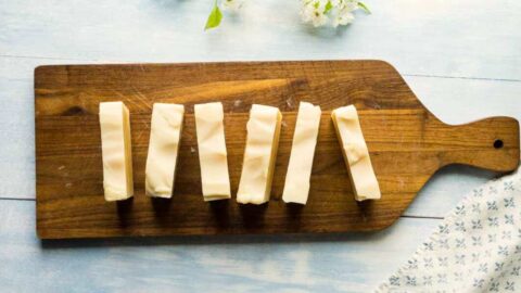 Image of homemade soap bars on a wooden board with a floral fabric and white blossoms on the side. The soaps have a creamy texture with slight ripples on the surface, suggesting a soft and natural product.