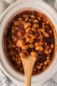 A close-up of a wooden spoon lifting a hearty scoop of barbecue beans from a slow cooker, showcasing the glossy sauce and the different types of beans merging together.