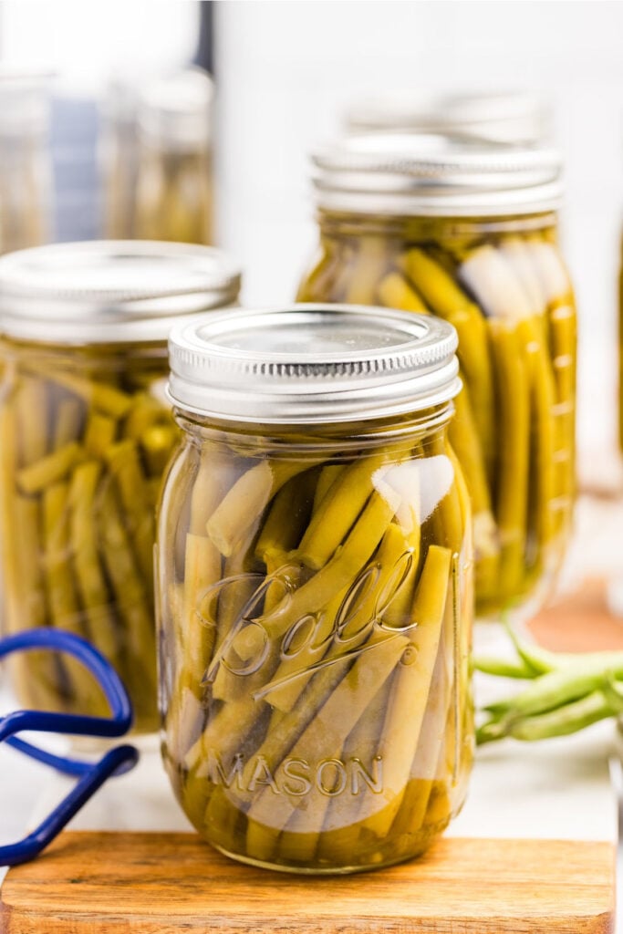 "A row of glass mason jars filled with pressure canned green beans, sealed with lids and placed on a kitchen counter next to a blue jar lifter."