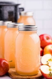 A quart-sized glass canning jar filled with homemade apple juice on a wooden cutting board, with fresh whole and halved apples to the side, suggesting a fresh, home-pressed juice.