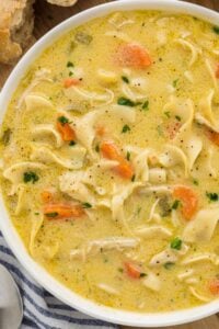 A creamy chicken noodle soup in a white bowl, garnished with parsley, with a visible chunk of bread beside the bowl.