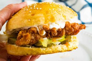 A close-up of a buffalo chicken sandwich showcasing the crispy chicken tender and fresh toppings of lettuce and pickles, with creamy dressing on a sesame seed bun.