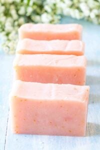 A row of homemade pale pink grapefruit soap bars lined up on a blue wooden surface with white flowers in the background.