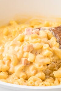 Creamy macaroni and cheese with ham served in a white bowl with a wooden spoon, emphasizing the dish's creamy consistency and generous mix of ham.